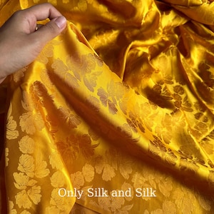 Könnte beinhalten: Nahaufnahme eines leuchtend gelben Seidenstoffs mit floralem Muster. Der Stoff ist drapiert und zeigt Falten und Schatten. Der Text "Only Silk and Silk" ist unten sichtbar. Der Stoff hat einen luxuriösen Glanz.
