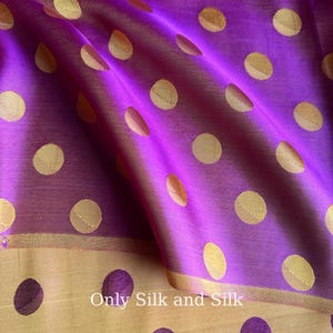 Könnte beinhalten: Nahaufnahme eines leuchtend lila Seidenstoffs mit großen, goldenen Polka-Dots. Der Stoff ist drapiert und zeigt einen goldenen Rand sowie den Text "Only Silk and Silk". Dieser Stoff eignet sich für Kleidung und Heimdekoration.