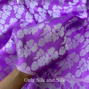 Könnte beinhalten: Nahaufnahme eines leuchtend lila Seidenstoffs mit einem Blumenmuster in einem helleren Lilaton. Der Stoff ist gefaltet und zeigt seine Textur und seinen Glanz. Der Text "Only Silk and Silk" ist unten sichtbar.