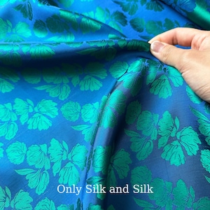 Könnte beinhalten: Nahaufnahme eines blauen und grünen floralen Seidenstoffs. Der Stoff hat ein sich wiederholendes Muster aus grünen Blumen auf blauem Grund. Der Text "Only Silk and Silk" ist auf dem Stoff zu sehen.