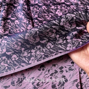 Può includere: Primo piano di un tessuto con motivo floreale. Il tessuto ha uno sfondo blu navy scuro con un motivo ripetuto di fiori viola chiaro. Il testo "Only Silk and Silk" è visibile.