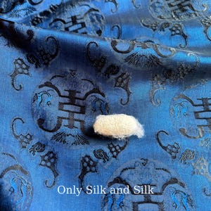 以下が含まれることがあります： 黒い装飾模様と白い蚕の繭が付いた青いシルク生地のクローズアップ。生地には光沢があり、下部には「Only Silk and Silk」というテキストがあります。