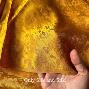 Könnte beinhalten: Nahaufnahme eines goldgelben Seidenstoffs mit floralem Muster. Der Stoff hat einen subtilen Glanz und ist drapiert, wodurch Falten und Texturen sichtbar werden. Der Text "Only Silk and Silk" ist unten sichtbar.