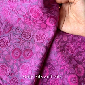 Könnte beinhalten: Nahaufnahme eines leuchtend magenta Seidenstoffs mit floralem Muster. Das Design zeigt detaillierte Rosen und andere Blumen, mit dem Text "Only Silk and Silk" sichtbar. Der Stoff hat einen luxuriösen Glanz.