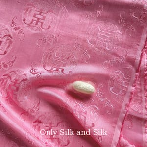 Könnte beinhalten: Rosa Seidenstoff mit sich wiederholendem Muster und dem Text "Only Silk and Silk". Ein weißer Seidenraupenkokon liegt auf dem Stoff. Der Stoff hat einen Glanz und eine subtile Textur.
