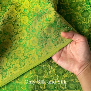 Può includere: Primo piano di un vibrante tessuto di seta verde con un motivo floreale in una tonalità più chiara di verde e oro. Il tessuto è piegato, rivelando la trama e il design. Il testo "Only Silk and Silk" è visibile.