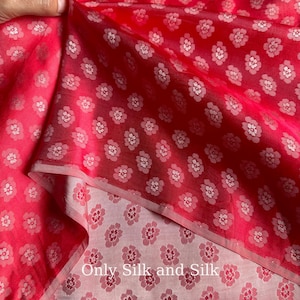 Könnte beinhalten: Nahaufnahme eines leuchtend roten Seidenstoffs mit einem zarten weißen Blumenmuster. Der Stoff ist gefaltet und zeigt eine hellere rosa Unterseite mit dem Text "Only Silk and Silk". Das Blumenmuster wiederholt sich über das Material.
