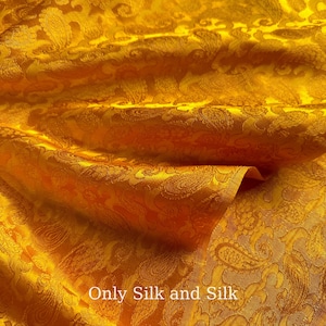 Könnte beinhalten: Nahaufnahme eines goldgelben Seidenstoffs mit Paisley-Muster. Der Stoff ist gefaltet und zeigt Textur und Glanz. Der Text "Only Silk and Silk" ist unten sichtbar.