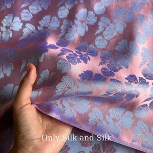 Könnte beinhalten: Nahaufnahme eines Stoffes mit floralem Muster. Der Stoff ist eine Mischung aus Rosa- und Lilatönen, wobei das florale Design in einem helleren Lilaton gehalten ist. Der Text "Only Silk and Silk" ist unten im Bild zu sehen.