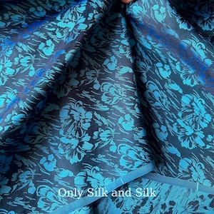 Può includere: Primo piano di un tessuto blu scuro con un motivo floreale nei toni del turchese e del blu. Il tessuto ha una lucentezza e il testo "Only Silk and Silk" è visibile. Probabilmente seta.