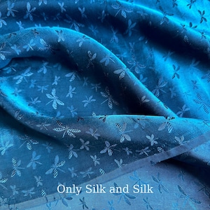 Könnte beinhalten: Nahaufnahme eines türkisfarbenen Seidenstoffs mit einem Libellenmuster. Der Stoff ist gefaltet, wodurch Schatten und Lichtreflexe entstehen, die die Textur und das Design hervorheben. Der Text "Only Silk and Silk" ist sichtbar.
