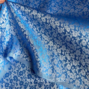 Könnte beinhalten: Nahaufnahme eines blauen Stoffes mit silbernem Blumenmuster. Der Stoff ist gefaltet und zeigt seine Textur und seinen Glanz. Der Text "Only Silk and Silk" ist unten im Bild zu sehen.