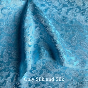 Könnte beinhalten: Nahaufnahme eines hellblauen Stoffes mit Paisley-Muster. Der Stoff hat einen dezenten Glanz und eine strukturierte Oberfläche. Der Text "Only Silk and Silk" ist unten im Bild zu sehen.