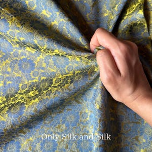 以下が含まれることがあります： 「Only Silk and Silk」のテキストが入った、青と黄色の花柄シルク生地。 手で生地を持ち、素材の質感とドレープを示しています。 花柄は細かく、複雑です。