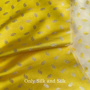 May include: Vibrant yellow silk fabric featuring a repeating pattern of silver floral motifs. The fabric is folded, highlighting its texture and lustre. The text "Only Silk and Silk" is displayed at the bottom of the image.