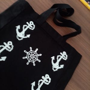 K&ouml;nnte beinhalten: Schwarze Stofftasche mit wei&szlig;en nautischen Motiven. Die Motive beinhalten Anker und ein Schiffssteuerrad.