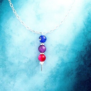 May include: A silver chain necklace with a pendant of three colorful beads: blue, purple, and red. The beads are spherical and arranged vertically. The background is a gradient of blue hues.