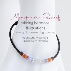 May include: A black cord bracelet featuring a central row of small beads in orange, purple, and translucent white, accented by silver-toned spacers and dark textured end beads. Text reads: Menopause Relief, calming hormonal fluctuations, energy + stamina + grounding, moonstone + carnelian, lapidiolite + hematite.