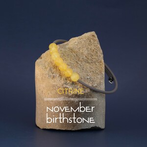 May include: Citrine birthstone bracelet with yellow beads on a gray cord. The bracelet is displayed on a rough stone with the text "CITRINE NOVEMBER BIRTHSTONE".