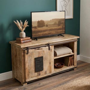 May include: A rustic wooden TV stand with sliding barn-style doors. The stand features a natural wood finish, a shelf, and a metal accent plate. A TV displays a landscape scene. Decorative items include a vase with dried plants and books.