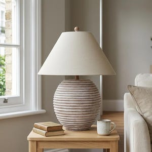 May include: A table lamp with a round, horizontally ribbed, light brown ceramic base and a tapered, off-white fabric lampshade. The lamp sits on a light oak wooden side table next to two small stacked books and a speckled ceramic mug.