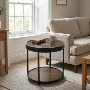 May include: A round side table with a black metal frame and two tiers. The top surface features a glass inlay with a decorative clock face design, while the lower shelf has a dark wood-grain finish. A small book and a ceramic mug sit on top.