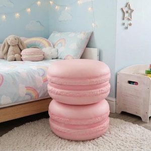 May include: Two-tiered pink macaron-shaped ottoman. The ottoman is in a child's bedroom with a bed featuring a rainbow and cloud-themed comforter. A stuffed bunny and pillows are on the bed. The room has a light blue wall.