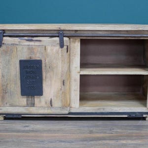 May include: A light-colored wooden cabinet with a sliding door and open shelving. The cabinet features a dark metal bar and hardware. A dark metal plaque on the door reads "Jupiter Iron Works Est 1925".