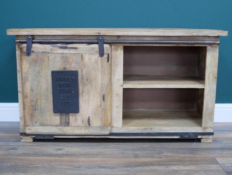 May include: A light-colored wooden cabinet with a sliding door and open shelving. The cabinet features a dark metal bar and hardware. A dark metal plaque on the door reads "Jupiter Iron Works Est 1925".