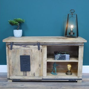 May include: A light-colored wooden cabinet with a sliding door and open shelving. The cabinet has a metal plate with text and is decorated with a small bonsai tree, a lamp, and other decorative items.