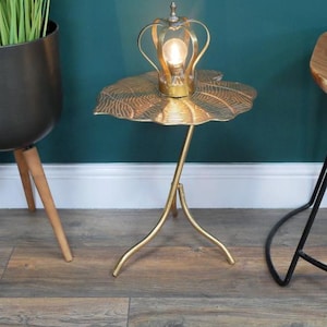 May include: A gold metal side table with a leaf-shaped top and a crown-shaped lamp. The table has three legs and is on a wooden floor.