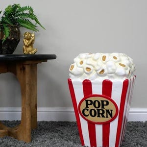 May include: A decorative popcorn-shaped container. The container is white with red vertical stripes and a red oval with the words "POP CORN". The top is filled with popcorn. A small gold monkey statue and a plant are on a table.