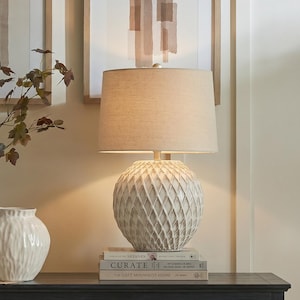 May include: A table lamp with a round, cream-coloured ceramic base featuring a raised, diamond-shaped lattice pattern. The lamp is topped with a cylindrical, light beige fabric shade. It rests on a stack of three books with visible text.
