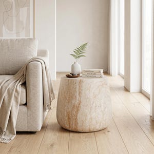 May include: A drum-shaped side table crafted from beige travertine stone with a natural marbled texture. The table surface holds a small white ceramic vase containing a green fern leaf and a stack of three books titled Natural Forms, Stone & Light.