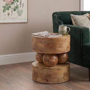 May include: A distinctive wooden side table with a cylindrical design. The table features two circular sections with three spherical accents. A magazine and a mug of coffee sit on top. A green sofa and framed botanical print are in the background.