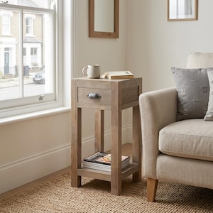 May include: A light brown wooden side table featuring a single drawer with a dark metal cup pull handle and an open lower shelf. The table surface holds a light-coloured ceramic mug and an open book, while the lower shelf contains a stack of magazines.