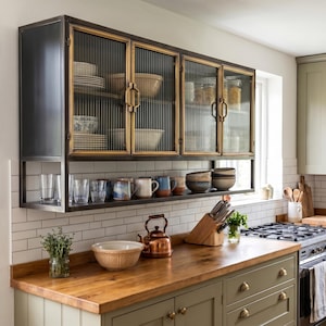 May include: A kitchen featuring a wooden worktop and olive green cabinets. Above, a black metal cabinet with glass doors displays crockery. A shelf below holds mugs and glasses. A copper kettle and bowl sit on the counter.