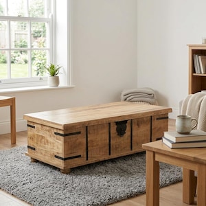 May include: A light brown wooden storage chest with black metal detailing. The chest, approximately 120 cm long, is on a grey rug. The room includes a window, a small table, and other wooden furniture.