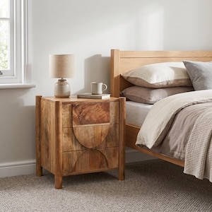May include: A light brown wooden bedside table with three drawers, each featuring a semi-circular carved detail. The table has four sturdy legs and a smooth finish, topped with a beige lamp and a white ceramic mug resting on a book.