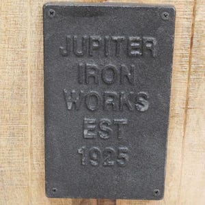 May include: A rectangular, dark gray metal plate with the embossed text "JUPITER IRON WORKS EST 1925" is affixed to a light-colored wooden surface. The plate is secured with four screws, one in each corner.