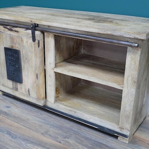 May include: A light-toned wooden sideboard featuring a sliding door on a dark metal rail system. The door has a rectangular black metal plaque with embossed text. The unit includes an open shelving section with two horizontal wooden shelves.