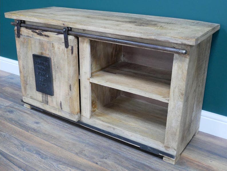 May include: A light-toned wooden sideboard featuring a sliding door on a dark metal rail system. The door has a rectangular black metal plaque with embossed text. The unit includes an open shelving section with two horizontal wooden shelves.