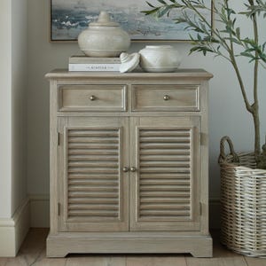 May include: A light oak cabinet with two drawers and slatted doors. The cabinet has a natural wood grain finish and brass-toned knobs. Decorative items sit on top, including a vase and a book.