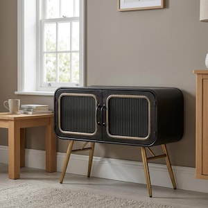 May include: Black cabinet with gold trim and legs. The cabinet has two doors with vertical ribbed detailing and gold trim. The legs are gold and angled. The cabinet is in a room with a window and a small wooden table.