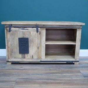 May include: A light-colored wooden cabinet with a sliding door and open shelving. The cabinet has a rectangular shape with a flat top and a dark metal accent on the door. The text "JUPITER IRON WORKS EST 1905" is on a metal plate on the door.