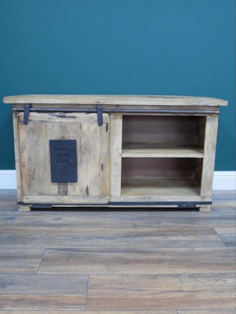 May include: A light-colored wooden cabinet with a sliding door and open shelving. The cabinet has a rectangular shape with a flat top and a dark metal accent on the door. The text "JUPITER IRON WORKS EST 1905" is on a metal plate on the door.