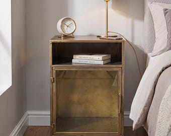May include: A rectangular brass-toned metal nightstand featuring an open top shelf and a lower cabinet with a glass-paneled door. The shelf holds two books titled The Night Read and Bedtime Tales, while a round clock sits on the top surface.