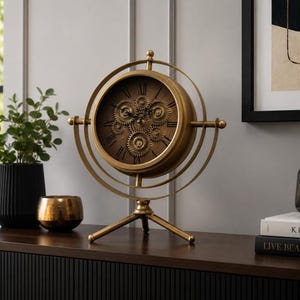 May include: A gold-toned metal table clock featuring a circular frame with a rotating gimbal design. The clock face displays Roman numerals and exposed mechanical gears in the centre. It is supported by a three-legged tripod base.