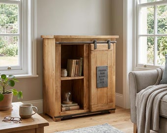 May include: A light brown wooden cabinet with a sliding door and open shelving. The cabinet has a metal bar and hardware. The door features a metal plaque that reads "Jupiter Iron Works Est 1876". Books and decorative items are visible inside.