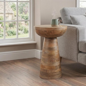 May include: A wooden side table with a round top and a conical base. The table is made of light brown wood and has a natural finish. A green mug sits on top of the table. The table is next to a grey sofa.